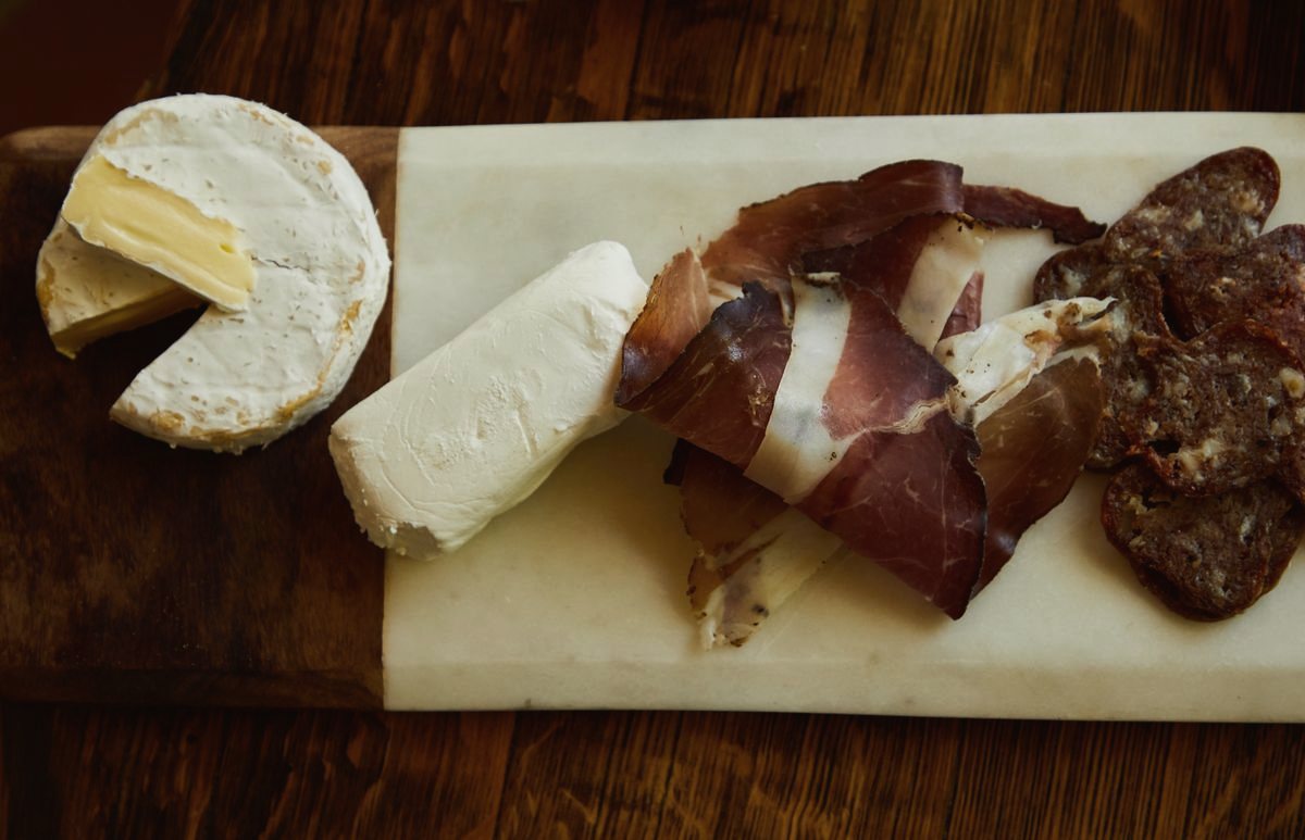 Brie, a log of soft white cheese, prosciutto, and salami on a marble board over dark wood